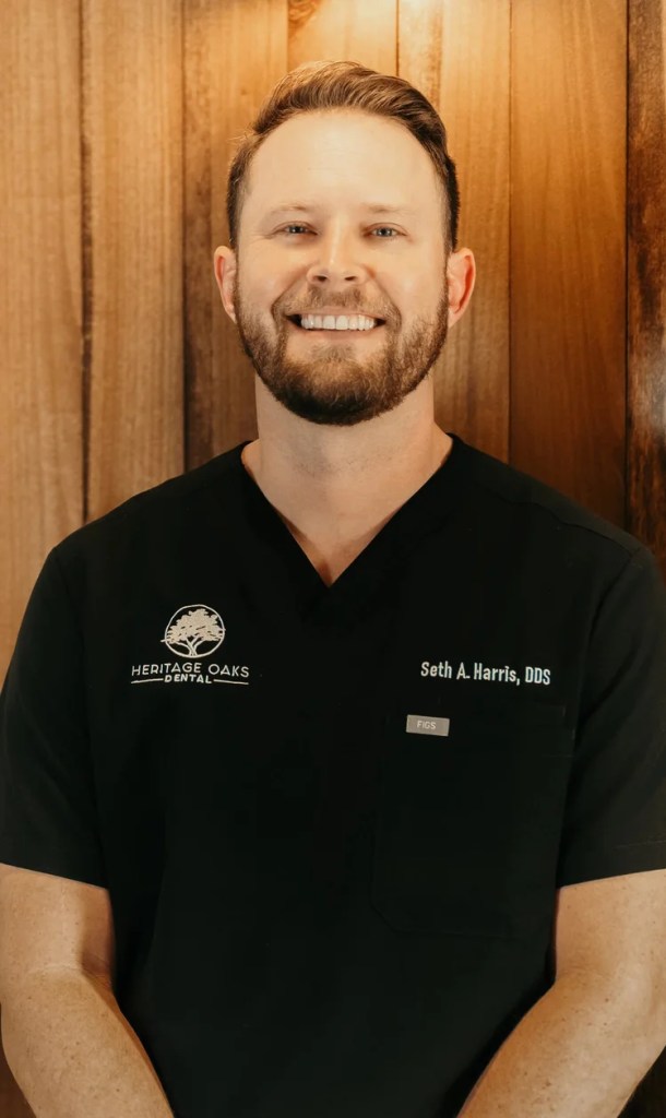 Professional headshot of Dr. Seth A. Harris smiling, wearing black medical scrubs against a warm wood panel backdrop.