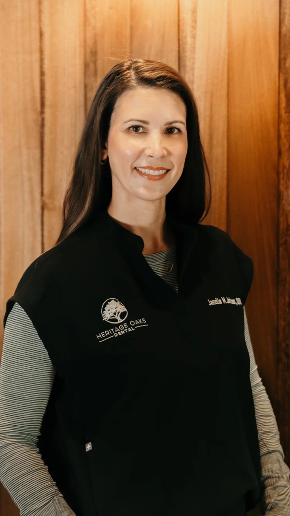 Professional headshot of Dr. Janelle M. Johnson smiling, wearing a black medical vest over a long-sleeved shirt against a warm wood panel backdrop.