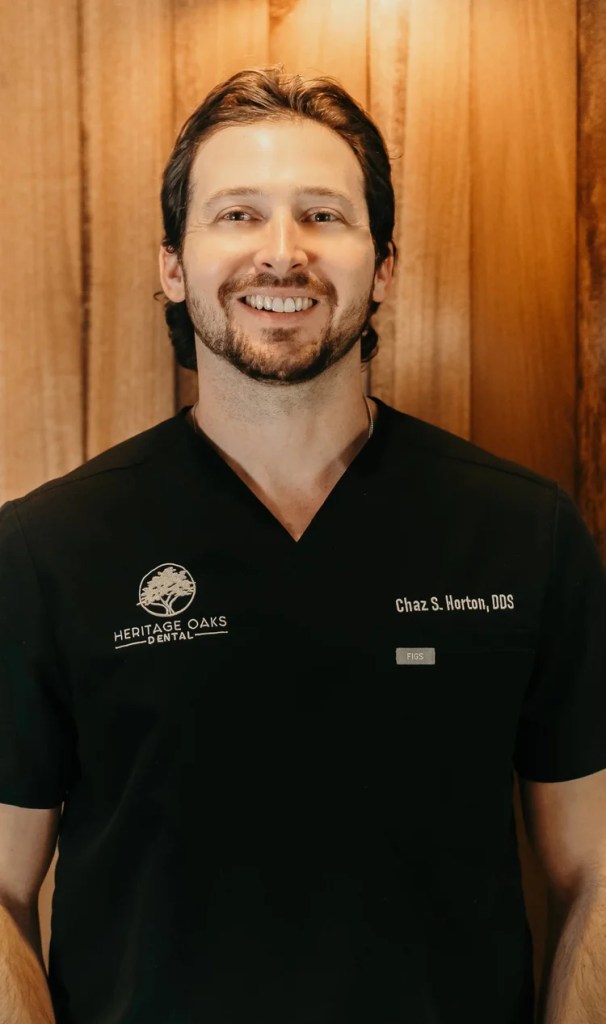 Professional headshot of Dr. Chaz S. Horton smiling, wearing black medical scrubs against a warm wood panel backdrop.